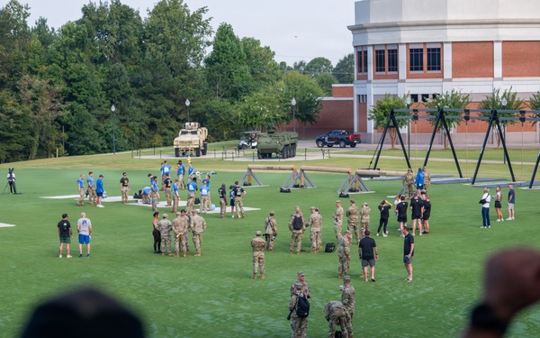 An outdoor event on a well-maintained green lawn appears to involve military personnel in camouflage uniforms interacting with civilians in casual and athletic wear, likely at a university or sports facility. Military equipment including what appears to be a tactical vehicle (MRAP) and training apparatus such as rope frames are visible on the field. Observers on an elevated platform or balcony are watching and photographing the event below.