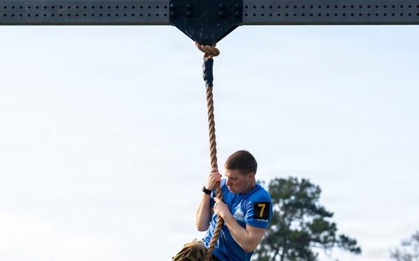 A male competitor wearing a blue shirt with the number '7' and camouflage pants is climbing a thick rope suspended from an overhead metal structure. The activity appears to be part of a military fitness competition or obstacle course challenge. Trees and an overcast sky are visible in the background.