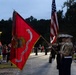 A U.S. Marine Corps color guard ceremony is taking place at MCB Camp Lejeune during early morning or dusk hours. Marines in dress uniforms are presenting the unit colors and the American flag to a gathered crowd of civilians and military personnel. The red unit guidon prominently displays the Marine Corps Eagle, Globe, and Anchor emblem with the inscription 'MCIEAST-MCB CAMP LEJEUNE'.