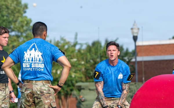 A group of military personnel are participating in the 'Best Squad Competition,' an Army fitness and skills event. Several soldiers wearing blue 'Best Squad Competition' t-shirts and camouflage pants are gathered around fitness equipment including a kettlebell and a blue barbell on a grassy outdoor area. A large red cylindrical obstacle is visible on the right side of the frame.