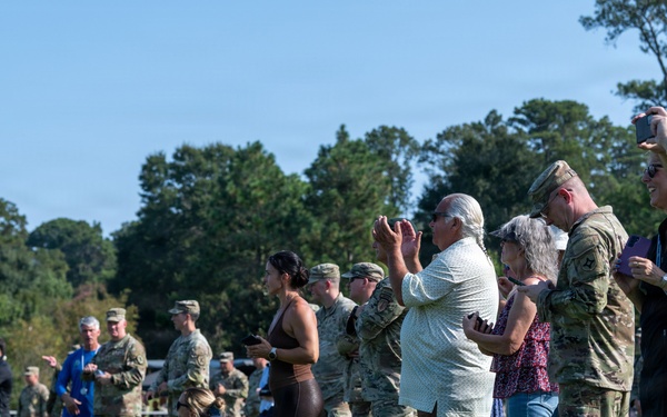 A military ceremony or event is taking place on a well-maintained grass field, with uniformed service members in camouflage (ACU/OCP pattern) standing alongside civilian family members or guests. Attendees are photographing and applauding, suggesting a recognition, promotion, or graduation ceremony. The setting appears to be a military installation or ROTC field during daytime under clear skies.