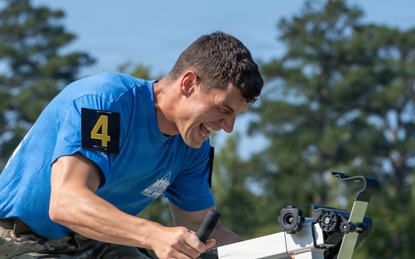 A man wearing a blue t-shirt with a yellow number '4' patch and military camouflage pants is intensely operating what appears to be a stationary exercise bike or ergometer outdoors. He is leaning forward with a strained, determined expression, suggesting he is in the middle of a competitive physical fitness event. The background features green trees and a clear blue sky.
