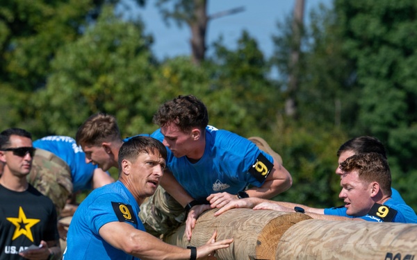 A group of individuals wearing blue team shirts numbered '9' are collaborating to lift or push a large wooden log mounted on a metal Rogue-branded rack, suggesting a military fitness competition or team challenge event. Personnel in U.S. Army uniforms and civilian attire observe in the background. The setting appears to be an outdoor athletic or training field with trees visible in the background.