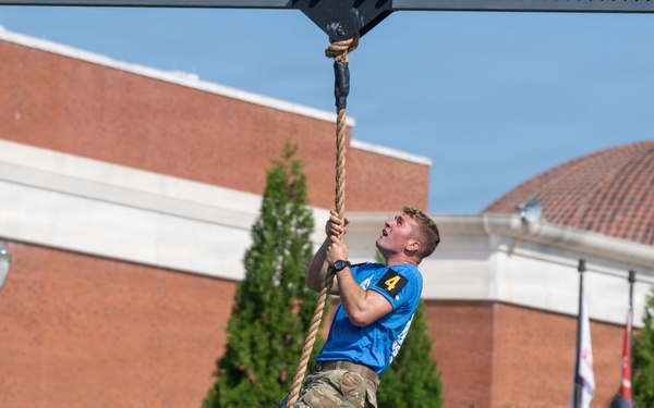A competitor wearing a blue jersey with the number 4 and military camouflage pants is climbing a thick rope suspended from a Rogue-branded rig structure. The event appears to be a military fitness competition held outdoors in front of a brick building, with flags and spectators visible in the background. The athlete is looking upward while gripping the rope with both hands and feet.