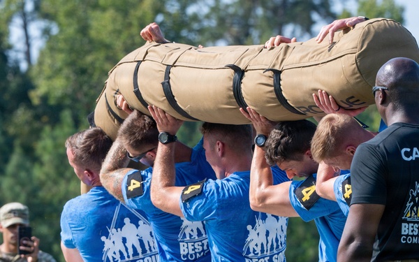 A group of military personnel in blue 'Best Squad Competition' t-shirts and camouflage pants are collectively lifting a large sandbag or log overhead during a team physical fitness event. A cadre member in a black 'Best Squad Competition' shirt observes from the right, while another uniformed soldier photographs the event in the background. The scene takes place outdoors with green trees visible in the background.