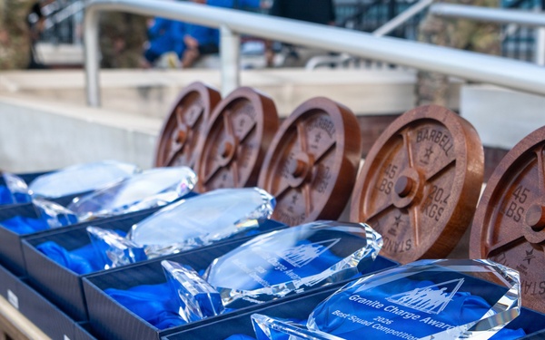 The image shows a display of awards arranged on a folding table, consisting of crystal glass trophies in blue-lined presentation boxes and decorative wooden barbell weight plate replicas. The crystal trophies are engraved and appear to be for the 'Granite Charge Award' for a 'Best Squad Competition.' A person in military camouflage uniform is partially visible on the left side of the frame.