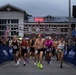 Runners are crossing the start line of the Marine Corps Half Marathon, held in front of the Wallace Creek Fitness Center. The race clock displays 00:00:00, indicating the race has just begun. A large crowd of participants and spectators is visible, with American flags and Marine Corps branding prominently displayed.