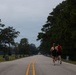 Two runners in athletic wear are jogging down the center of a straight, two-lane road lined with small memorial or commemorative signs on both sides. The road is flanked by tall pine and deciduous trees under an overcast sky. The perspective creates a strong vanishing point, emphasizing the length of the road ahead.