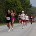A road race is underway on a paved two-lane road lined with trees. Multiple runners of varying ages and fitness levels are competing, with the foreground runner wearing bib number 659. The event appears to be an organized race, likely a 5K or similar community run, with spectators visible along the roadside in the background.