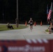 A runner wearing bib number 894 approaches the finish line of what appears to be a road race, likely a 5K or similar event. Spectators line both sides of the course, with orange traffic cones marking the route and flags including the American flag visible in the background. A finish line banner with 'Marine Corps' text is partially visible and blurred in the foreground.