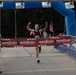 A runner wearing bib number 894 and a Marines singlet crosses the finish line of the Marine Corps Half Marathon with arms raised in victory. Two uniformed Marines hold the finish line tape on either side of the inflatable blue arch. The event appears to take place outdoors on a paved surface with spectators and flags visible in the background.