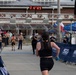 A female runner in black athletic wear is approaching the finish line of the Marine Corps Half Marathon. The race clock overhead displays a time of 1:27:29. Spectators and race officials line the finishing chute as the runner nears completion of the event.