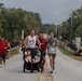A road race event is taking place on a tree-lined street that crosses a bridge over a waterway. Several runners are visible, with the foreground featuring a man pushing a double jogging stroller containing two young children, accompanied by two other runners wearing race bibs numbered 1273 and 1274. The overcast sky and lush green surroundings suggest a warm, humid climate, likely in the southeastern United States.