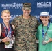 Three men pose together at the Marine Corps Half Marathon event, held at Camp Lejeune-New River. Two runners holding crystal/glass trophies and wearing race medals flank a U.S. Marines officer in camouflage uniform. The setting appears to be an awards ceremony following the race.