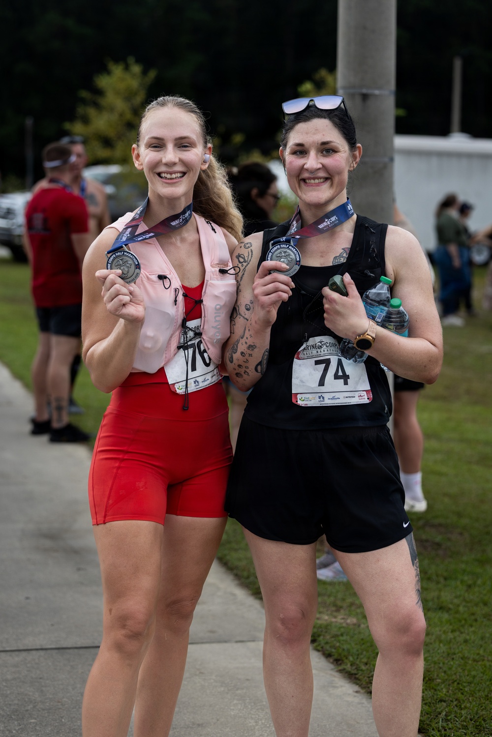 Two women pose together after completing the Marine Corps Half Marathon, each displaying their finisher medals. The woman on the left wears red athletic wear with bib number 76, while the woman on the right wears black athletic wear with bib number 74. Both appear sweaty and elated, suggesting they have just finished the race.