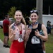 Two women pose together after completing the Marine Corps Half Marathon, each displaying their finisher medals. The woman on the left wears red athletic wear with bib number 76, while the woman on the right wears black athletic wear with bib number 74. Both appear sweaty and elated, suggesting they have just finished the race.