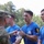 2026 US Army Best Squad Competition: Community Day Fitness Event A military award ceremony is taking place outdoors, where soldiers in blue 'Best Squad Competition' t-shirts are receiving a crystal trophy from a uniformed Army officer in camouflage. The recipients are wearing camouflage pants and numbered armbands, indicating they are competition participants. The setting appears to be a grassy outdoor area with trees in the background.