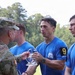 A military award ceremony is taking place outdoors, where soldiers in blue 'Best Squad Competition' t-shirts are receiving a crystal trophy from a uniformed Army officer in camouflage. The recipients are wearing camouflage pants and numbered armbands, indicating they are competition participants. The setting appears to be a grassy outdoor area with trees in the background.