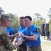 A uniformed U.S. Army soldier is presenting a crystal/glass trophy award to competitors wearing blue 'Best Squad Competition' t-shirts with number '9' armbands. The ceremony appears to be an outdoor award presentation following a military squad competition. At least three competitors in blue shirts are visible receiving the award.