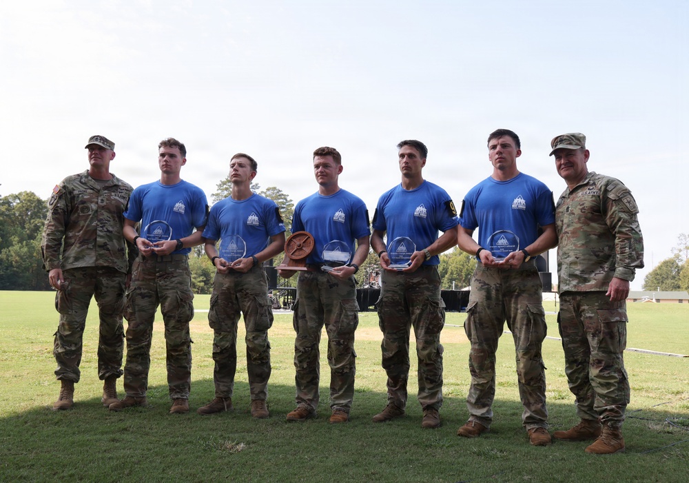 A group of seven military personnel pose for a formal photograph on a grass field. Five individuals in the center wear matching blue event t-shirts and hold clear acrylic awards and one wooden trophy, suggesting a competition or ceremony recognition. Two soldiers in full camouflage uniforms flank the group on either side, likely serving as presiding officers.