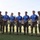2026 US Army Best Squad Competition: Community Day Fitness Event A group of seven military personnel pose for a formal photograph on a grass field. Five individuals in the center wear matching blue event t-shirts and hold clear acrylic awards and one wooden trophy, suggesting a competition or ceremony recognition. Two soldiers in full camouflage uniforms flank the group on either side, likely serving as presiding officers.