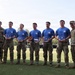 A group of seven military personnel pose for a formal photograph on a grass field. Five individuals in the center wear matching blue event t-shirts and hold clear acrylic awards and one wooden trophy, suggesting a competition or ceremony recognition. Two soldiers in full camouflage uniforms flank the group on either side, likely serving as presiding officers.