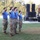 2026 US Army Best Squad Competition: Community Day Fitness Event Four military personnel wearing blue t-shirts and camouflage pants are standing at attention and saluting on a grass field during what appears to be an outdoor military ceremony or event. A uniformed officer is speaking at a podium on a stage in the background, with large speaker stacks visible. The setting appears to be a formal military gathering held outdoors during daylight hours.