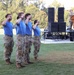 Four military personnel wearing blue t-shirts and camouflage pants are standing at attention and saluting on a grass field during what appears to be an outdoor military ceremony or event. A uniformed officer is speaking at a podium on a stage in the background, with large speaker stacks visible. The setting appears to be a formal military gathering held outdoors during daylight hours.