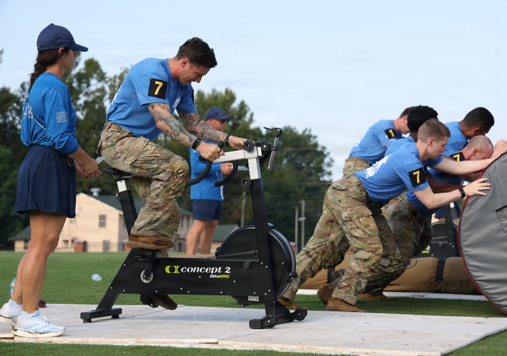 Military personnel in camouflage uniforms and blue shirts are participating in a fitness competition or team challenge event outdoors on a turf field. One competitor wearing number 7 is pedaling a Concept2 BikeERG stationary bike while others in the background appear to be pushing or moving a large object, likely a tent or heavy bag. A female observer in a blue cap and skirt monitors the activity nearby.