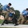 2026 US Army Best Squad Competition: Community Day Fitness Event Military personnel in camouflage uniforms and blue shirts are participating in a fitness competition or team challenge event outdoors on a turf field. One competitor wearing number 7 is pedaling a Concept2 BikeERG stationary bike while others in the background appear to be pushing or moving a large object, likely a tent or heavy bag. A female observer in a blue cap and skirt monitors the activity nearby.