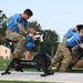 Military personnel in camouflage uniforms and blue shirts are participating in a fitness competition or team challenge event outdoors on a turf field. One competitor wearing number 7 is pedaling a Concept2 BikeERG stationary bike while others in the background appear to be pushing or moving a large object, likely a tent or heavy bag. A female observer in a blue cap and skirt monitors the activity nearby.