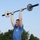 2026 US Army Best Squad Competition: Community Day Fitness Event A male athlete wearing military camouflage pants and a blue t-shirt is performing an overhead barbell press outdoors. The barbell is a Rogue-branded bar with white bumper plates loaded on each end. The setting appears to be a military fitness competition held on a grassy field.