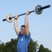 A male athlete wearing military camouflage pants and a blue t-shirt is performing an overhead barbell press outdoors. The barbell is a Rogue-branded bar with white bumper plates loaded on each end. The setting appears to be a military fitness competition held on a grassy field.