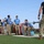2026 US Army Best Squad Competition: Community Day Fitness Event Military personnel in blue t-shirts and camouflage pants participate in the 'Best Squad Competition,' an organized fitness and strength event. Competitors are performing barbell lifts while cadre members in black 'Best Squad Competition' shirts observe and supervise. The event appears to take place on a grassy outdoor area of a military installation.