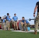 Military personnel in blue t-shirts and camouflage pants participate in the 'Best Squad Competition,' an organized fitness and strength event. Competitors are performing barbell lifts while cadre members in black 'Best Squad Competition' shirts observe and supervise. The event appears to take place on a grassy outdoor area of a military installation.