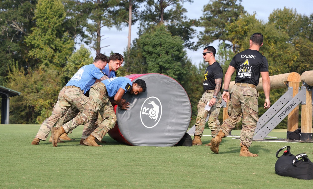 A group of military personnel in camouflage uniforms and blue shirts are pushing a large padded cylindrical roller across a grass field during what appears to be the Army Best Squad Competition. Two cadre members in black shirts observe and oversee the activity. The event appears to be a team physical fitness or obstacle course challenge at a military training facility.
