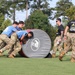A group of military personnel in camouflage uniforms and blue shirts are pushing a large padded cylindrical roller across a grass field during what appears to be the Army Best Squad Competition. Two cadre members in black shirts observe and oversee the activity. The event appears to be a team physical fitness or obstacle course challenge at a military training facility.