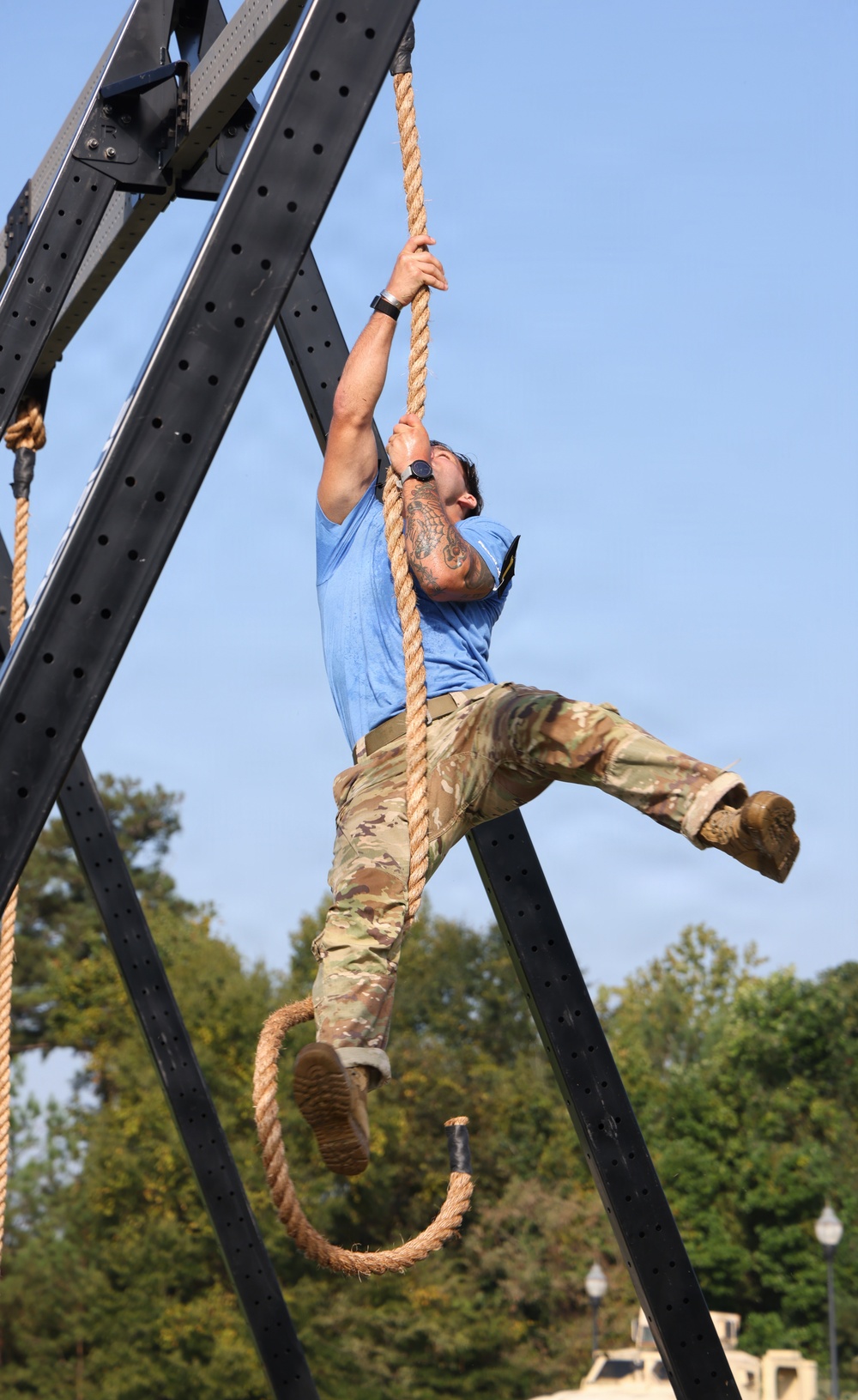 A person is climbing a thick rope suspended from a large black metal A-frame structure outdoors. The individual is wearing a blue t-shirt, military-pattern camouflage trousers, and combat boots, and has visible tattoos on one arm. The setting appears to be a military or fitness obstacle course, with trees and what appears to be military vehicles visible in the background.