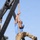 2026 US Army Best Squad Competition: Community Day Fitness Event A person is climbing a thick rope suspended from a large black metal A-frame structure outdoors. The individual is wearing a blue t-shirt, military-pattern camouflage trousers, and combat boots, and has visible tattoos on one arm. The setting appears to be a military or fitness obstacle course, with trees and what appears to be military vehicles visible in the background.