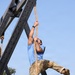 A person is climbing a thick rope suspended from a large black metal A-frame structure outdoors. The individual is wearing a blue t-shirt, military-pattern camouflage trousers, and combat boots, and has visible tattoos on one arm. The setting appears to be a military or fitness obstacle course, with trees and what appears to be military vehicles visible in the background.