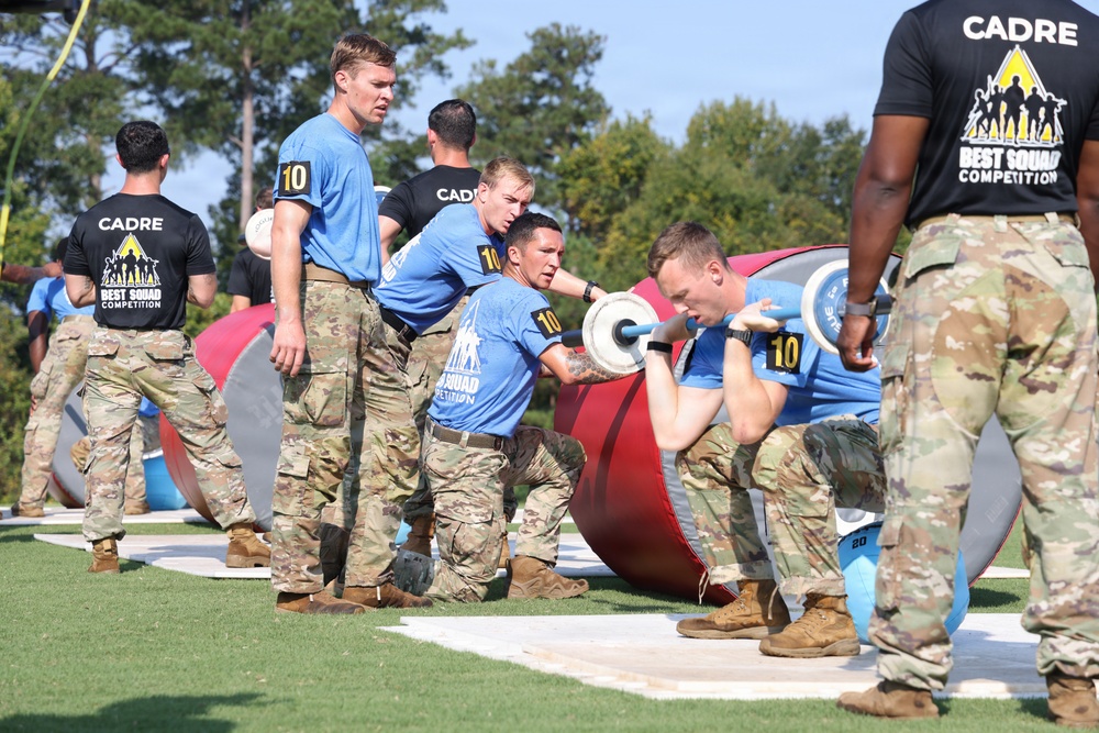 Military personnel are competing in what appears to be the 'Best Squad Competition,' an organized military fitness event. Team members wearing blue shirts with the number '10' are performing a barbell lift exercise while cadre members in black shirts observe and supervise. The event takes place outdoors on a grass/turf field with various large cylindrical obstacles visible in the background.