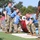 2026 US Army Best Squad Competition: Community Day Fitness Event Military personnel are competing in what appears to be the 'Best Squad Competition,' an organized military fitness event. Team members wearing blue shirts with the number '10' are performing a barbell lift exercise while cadre members in black shirts observe and supervise. The event takes place outdoors on a grass/turf field with various large cylindrical obstacles visible in the background.