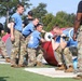 Military personnel are competing in what appears to be the 'Best Squad Competition,' an organized military fitness event. Team members wearing blue shirts with the number '10' are performing a barbell lift exercise while cadre members in black shirts observe and supervise. The event takes place outdoors on a grass/turf field with various large cylindrical obstacles visible in the background.