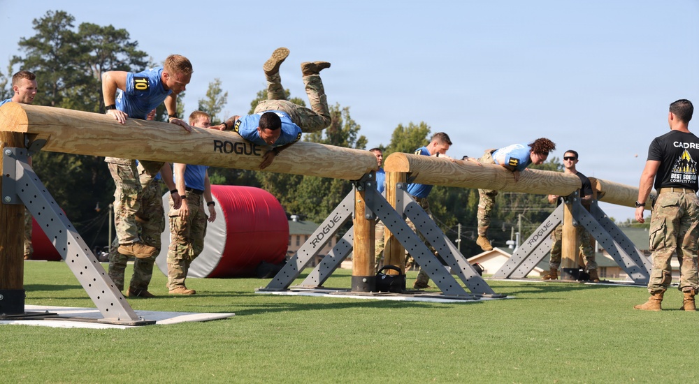 Military personnel are competing in an obstacle course challenge involving large horizontal logs mounted on Rogue-branded wooden supports. Competitors wearing blue athletic shirts are climbing over or traversing the log obstacles while others in military camouflage uniforms observe or assist. A person wearing a black 'CADRE' shirt on the right appears to be officiating or supervising the competition.