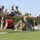 2026 US Army Best Squad Competition: Community Day Fitness Event Military personnel are competing in an obstacle course challenge involving large horizontal logs mounted on Rogue-branded wooden supports. Competitors wearing blue athletic shirts are climbing over or traversing the log obstacles while others in military camouflage uniforms observe or assist. A person wearing a black 'CADRE' shirt on the right appears to be officiating or supervising the competition.