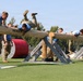 Military personnel are competing in an obstacle course challenge involving large horizontal logs mounted on Rogue-branded wooden supports. Competitors wearing blue athletic shirts are climbing over or traversing the log obstacles while others in military camouflage uniforms observe or assist. A person wearing a black 'CADRE' shirt on the right appears to be officiating or supervising the competition.