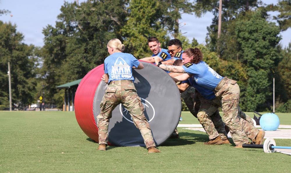 A group of four individuals wearing blue team shirts and military camouflage pants are collaboratively pushing a large oversized ball during what appears to be a military fitness competition or team challenge event. The participants are working together on a grassy field, with additional equipment such as a barbell and another ball visible in the background. The event appears to be an organized outdoor military-style athletic competition.
