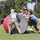 2026 US Army Best Squad Competition: Community Day Fitness Event A group of four individuals wearing blue team shirts and military camouflage pants are collaboratively pushing a large oversized ball during what appears to be a military fitness competition or team challenge event. The participants are working together on a grassy field, with additional equipment such as a barbell and another ball visible in the background. The event appears to be an organized outdoor military-style athletic competition.