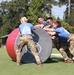 A group of four individuals wearing blue team shirts and military camouflage pants are collaboratively pushing a large oversized ball during what appears to be a military fitness competition or team challenge event. The participants are working together on a grassy field, with additional equipment such as a barbell and another ball visible in the background. The event appears to be an organized outdoor military-style athletic competition.