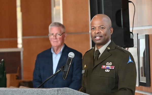 A military officer in dress uniform stands at a podium delivering a speech at what appears to be an official ceremony or event. The podium displays a distinctive triangular logo in blue and green, consistent with U.S. Army Corps of Engineers or a similar military organization. A civilian man in a dark suit stands in the background.